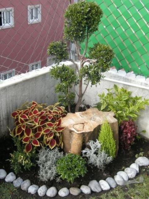 a small corner garden surrounded with pebbles, with bright plants, a small tree and a tree stump is a lovely idea for a small space