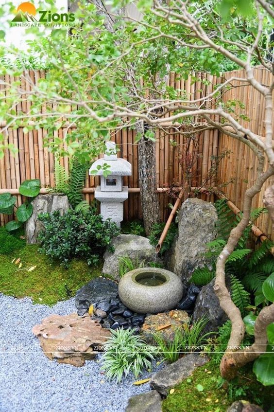 a small Japanese corner garden with rocks, a bamboo fountain, greenery, moss and trees and a stone lantern