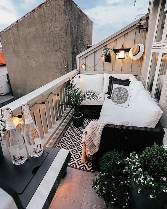 a small Scandinavian balcony with a printed rug, a wicker corner sofa, potted plants and blooms and some lights