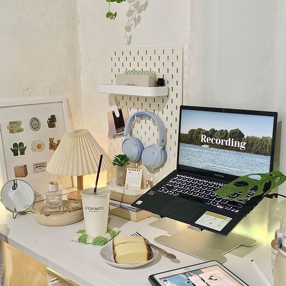 a small study space with a laptop on a stand, a memo board, a pegboard with stuff, some decor, lamps and food