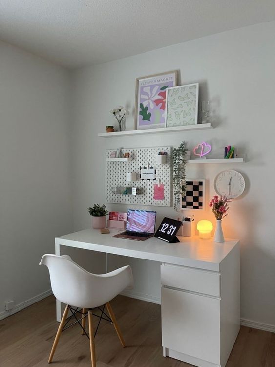 a small white workspace with a desk and a chair, a laptop and a clock, a pegboard for holding stuff, shelves with decor and a table lamp