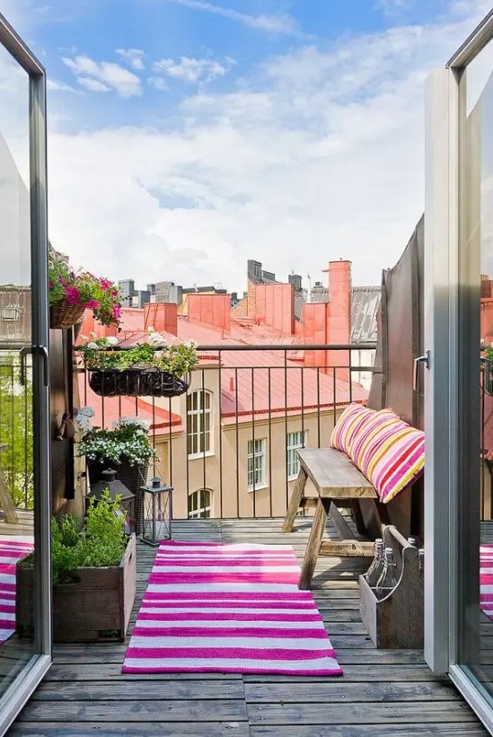 a small yet bright summer balcony with with a simple wooden bench, colorful striped textiles, potted greenery and blooms to catch some sun