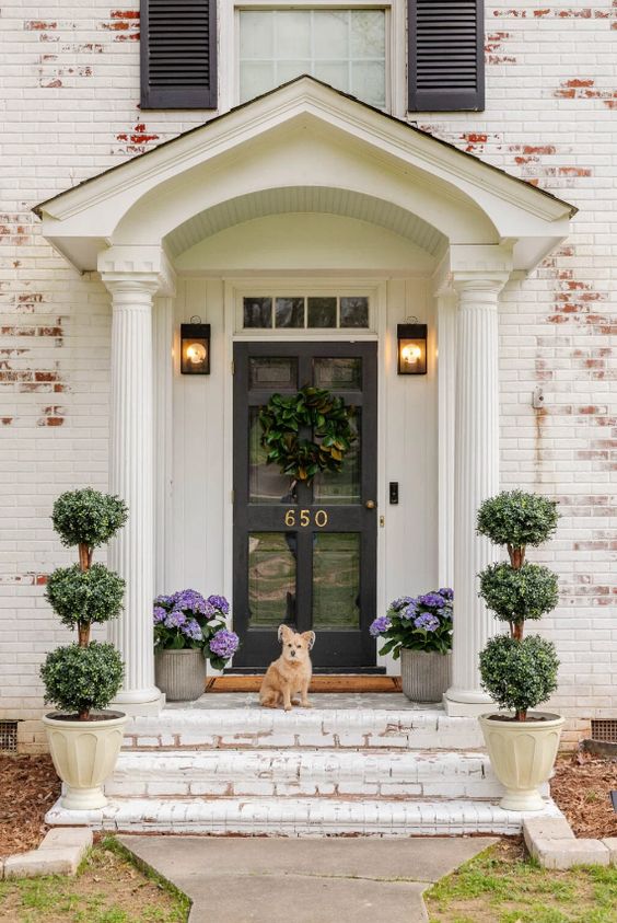 a sophisticated and chic front porch with cool lamps, a glass pane door, potted hydrangeas and topiaries is a cool space