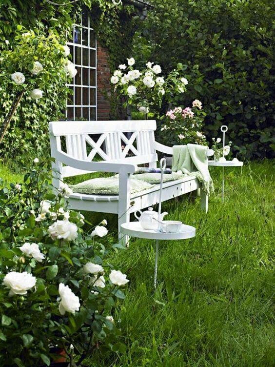 a stunning cottage garden nook with a green lawn, blooming bushes, white furniture and a white sofa with some green textiles