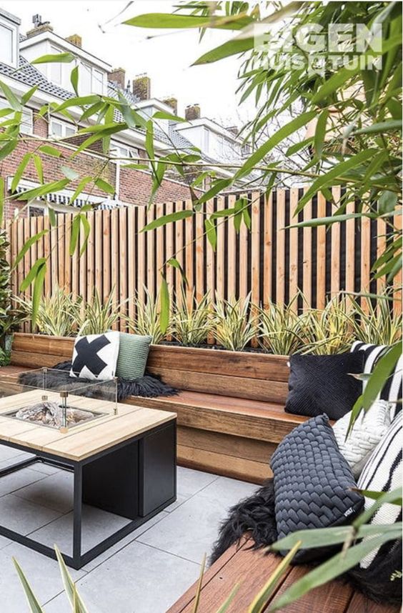 a stylish modern terrace with a light-stained fence and a planter with greenery, stained benches and a fire pit