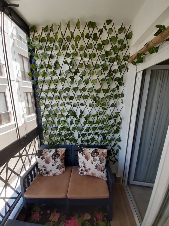 a tiny balcony with a loveseat, a trellis covered with faux leaves used to make the space feel more outdoorsy and cool