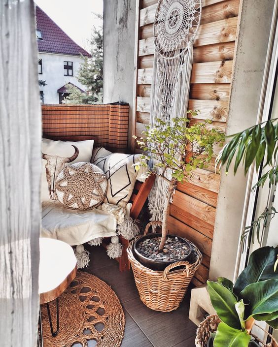 a tiny boho balcony with a bench with boho pillows and blankets, potted plants and greenery, a dream catcher, a jute rug and a wood slice table