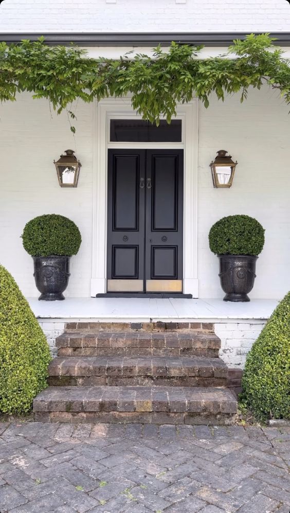 a vintage-inspired porch with a black door, vintage lamps on the walls, greenery over the entrance and green topiaries