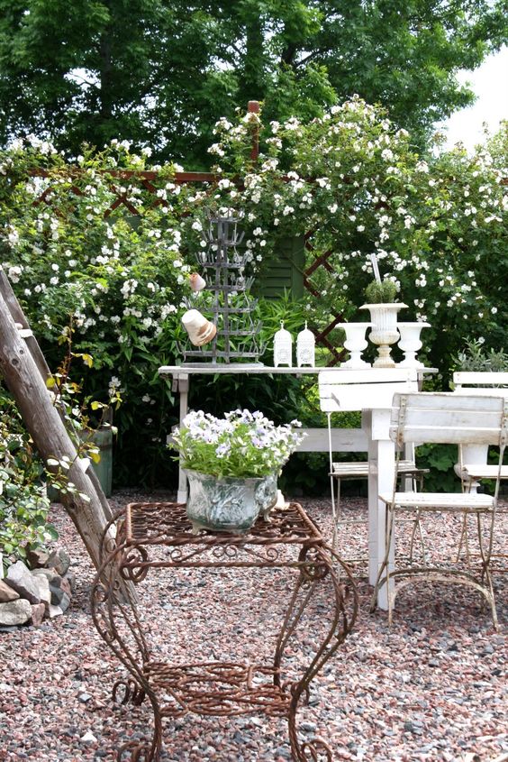 a vintage secret garden with blooming bushes, a gravel terrace, white garden furniture, some urns and pots for gardening