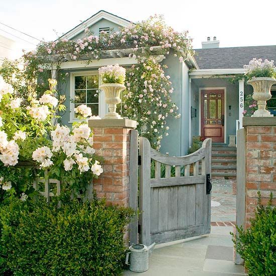 a weathered wood trellis covered with white blooms and greenery, a simple matching gate and lush flowers climbing up the facade of the house