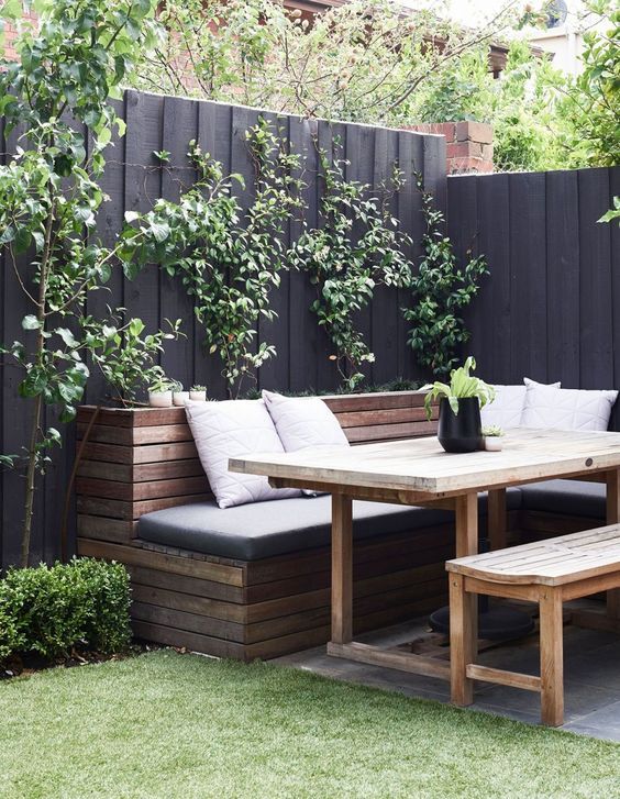 a welcoming corner with a black wooden fence, stained benches and a table, a trellis with greenery that refreshes the space