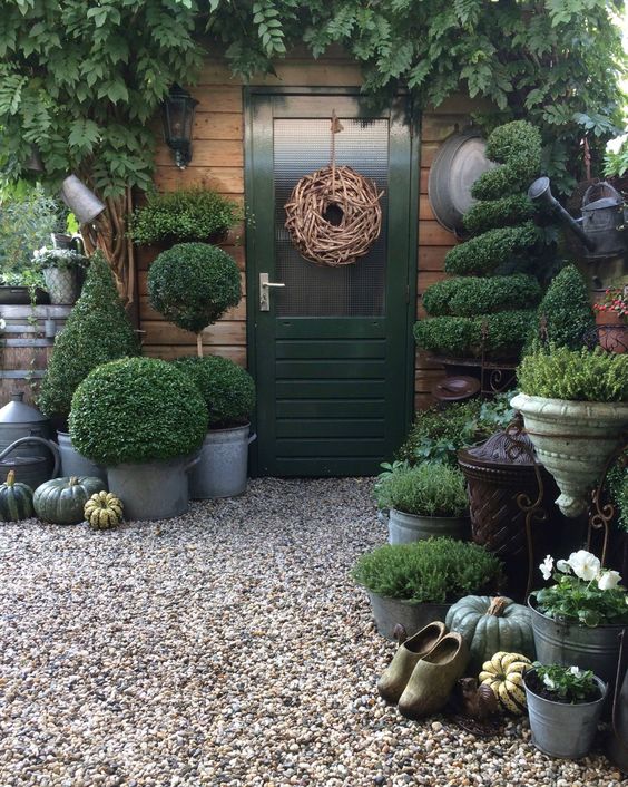 a whimsical entrance with a green door and a vine wreath, greenery over the door and lots of various topiaries and greenery in pots