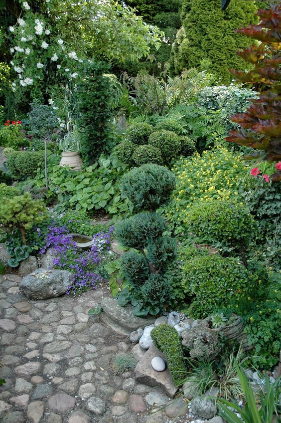 a whimsical shade garden with greenery, bold blooms, shrubs, blooming trees and usual ones, a pebble path