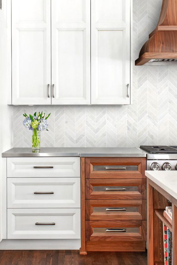 a white and stained kitchen with white and metal countertops, a grey marble chevron backsplash and a stained hood over the cooker