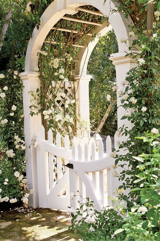 a white gate with a large arbor over it, with greenery and white blooming vines covering the arbor  is adorable