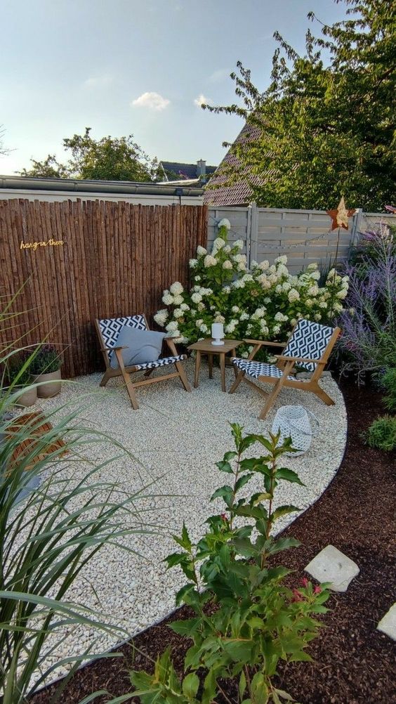 a white gravel patio with pritned chairs, a side table, some blooms and greenery is a cool idea to style the space by the fence