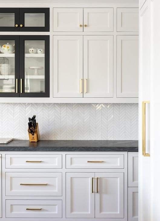 a white kitchen with shaker cabinets, grey countertops, a white chevron tile backsplash and gold fixtures