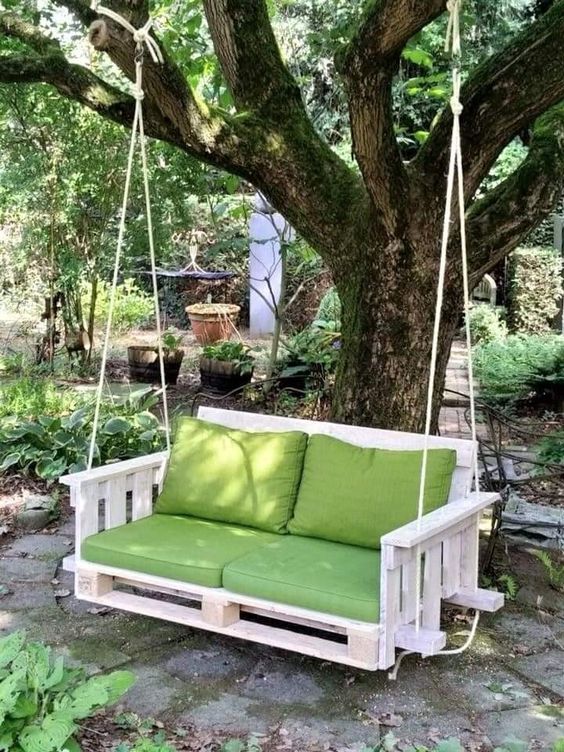 a DIY pallet chair hanging on a tree, with green cushions that match the greenery around is a great cottage-inspired item
