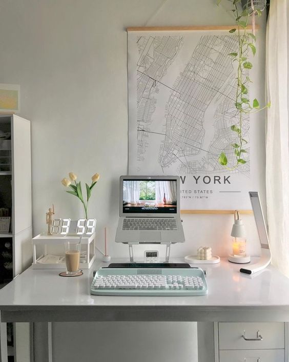 a white workspace with a laptop on a stand, a green keyboard, a clock and some blooms on a stand and map wall decor