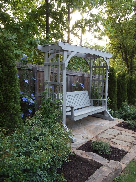 a whitewashed wooden arbor with a bench, surrounded with greenery and blooms is put in a shady corner to avoid the sun