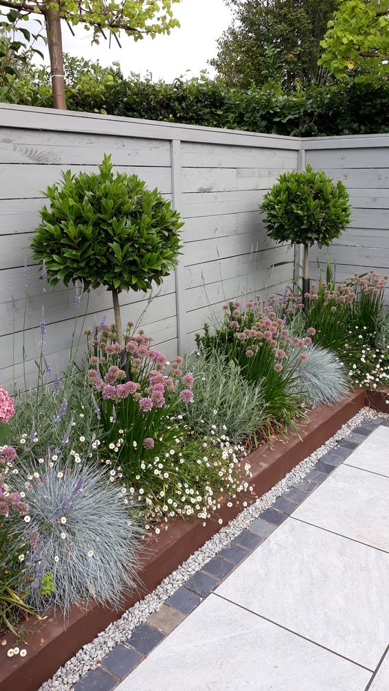 a whitewashed wooden fence with a raised garden bed with greenery, blooms and topiary trees are a chic and cool combo