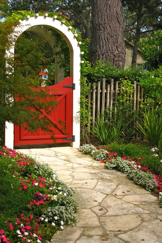 a wood fence, a red gate and a white arbor plus greenery covering it, bold red and white blooms lining up the stone garden path