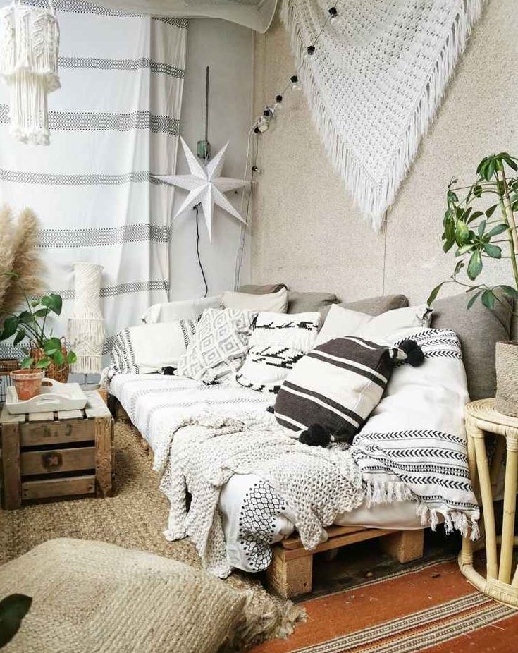 a boho balcony with a jute rug and cushions, a pallet sofa with black and white boho pillows, macrame and blankets, a crate as a table and some potted plants