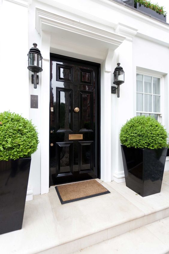 an elegant and refined entrance with a glossy black door, tall black planters with greenery and vintage wall lamps