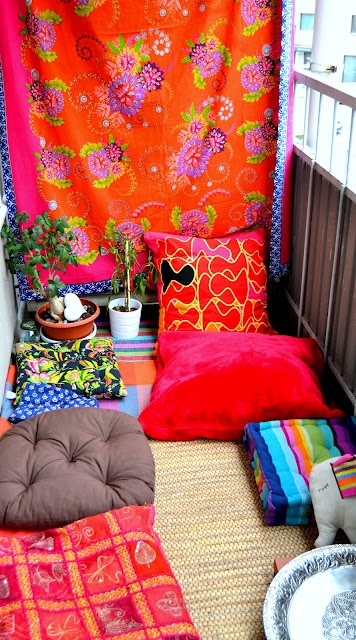 a colorful boho balcony with an orange curtain, super bright and printed pillows, a jute rug, potted greenery, a striped bold rug