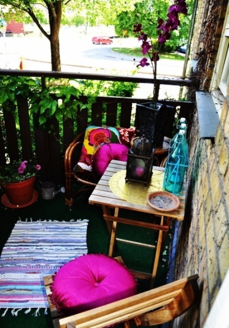 a super colorful boho balcony with wood and rattan furniture, bright pink pillows and a colorful rug, a side table with Moroccan lanterns and potted blooms