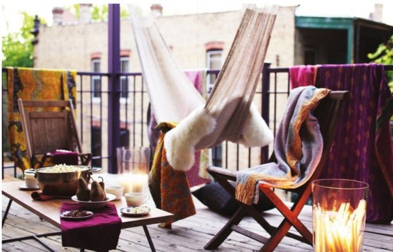 a colorful boho balcony with folding chairs with bright covers, a table, a hammock with lots of pillows and blankets and a large candle holder is very inviting