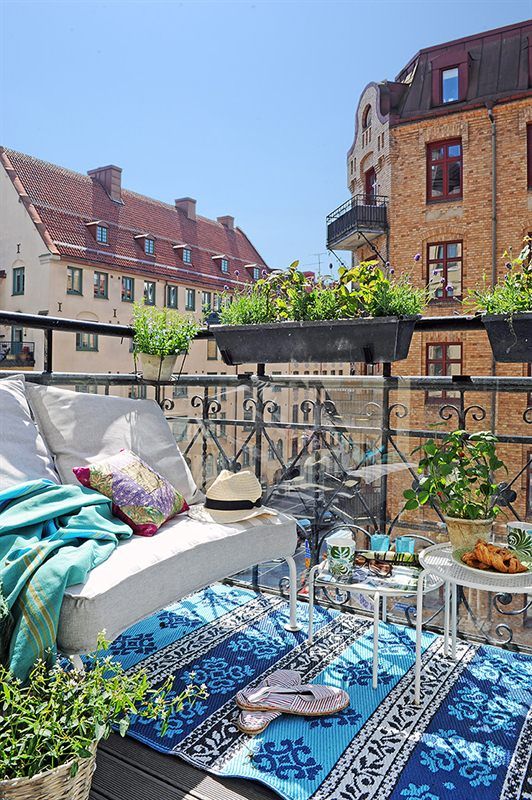 a boho-inspired balcony wiht a white loveseat with cozy pillows and a blanket, a bold printed blue rug, side tables and lots of potted greenery around