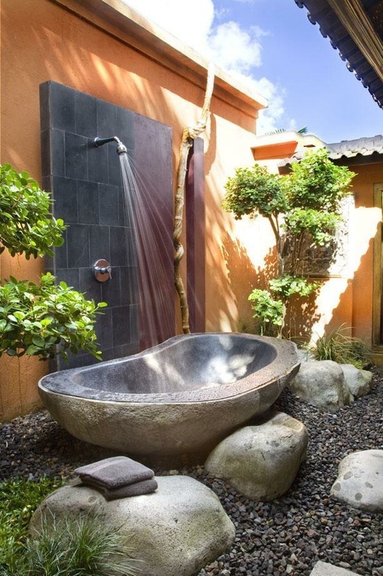 an outdoor tropical bathroom with a metal tub surrounded with rocks and a shower right here plus beautiful mini trees around