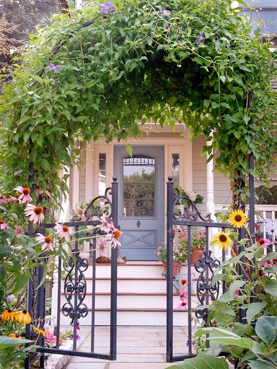 A beautiful entrance with a metal gate and an arch over it covered with greenery, bold blooms and a cozy cottage porch with a grey door. 