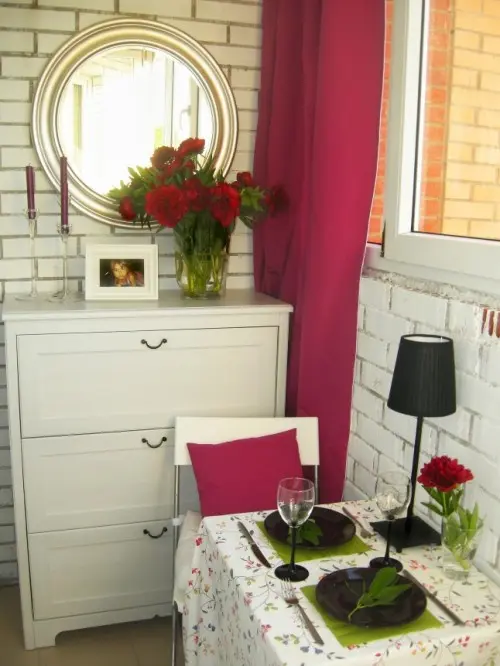 a small balcony turned into a mini living room with a dresser and a mirror, a table with a couple of chairs, bold curtains and a table lamp