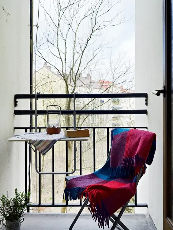 a tiny balcony with a folding table and a chair, some greenery and a blanket that will make your feel comfy and cozy in fall and winter