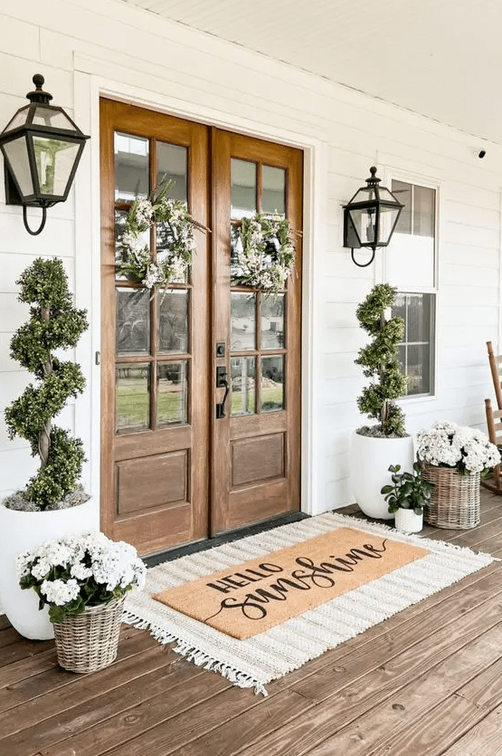 a beautiful spring farmhouse porch with layered rugs, potted blooms and greenery, greenery and white flower wreaths