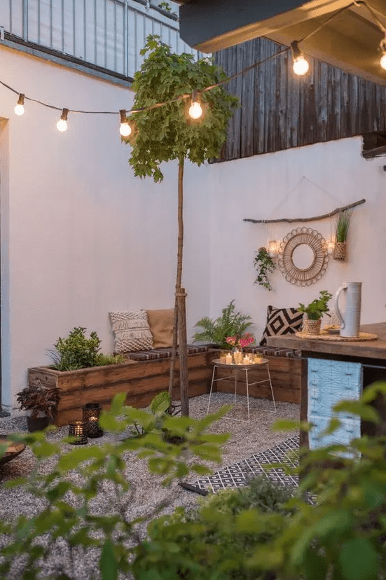 a boho patio done with gravel, with a stained corner bench and printed pillows and cushions, some boho decor and greenery