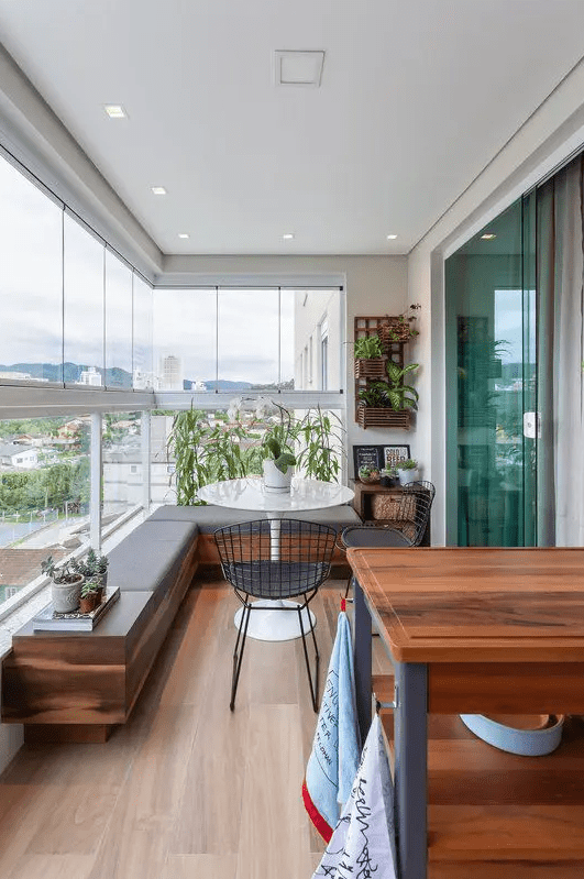 a chic modern balcony with a built-in upholstered bench, metal chairs, a round table, potted greenery, a cart for cooking or serving drinks