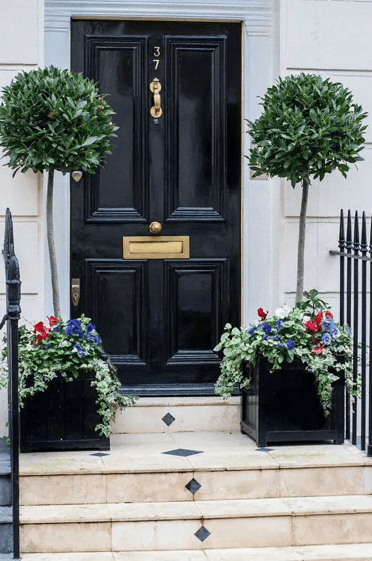 a chic small porch with a black door and bay tree topiaries with flower underplantings that give this front door a chic look