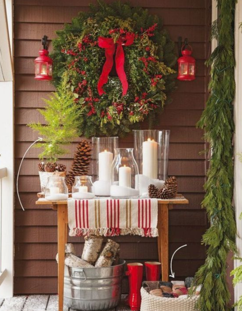 a console table with pillar candles, pinecones, an evergreen wreath with red berries and bows, red lanterns and firewood in a bucket