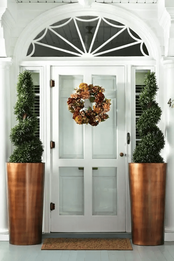 a little porch with a white front door with glass panes and tall copper planters with creative topiary trees