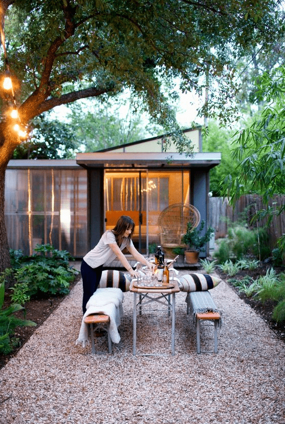 a modern backyard with greenery and trees, wooden and metal benches and a dining table plus striped pillows