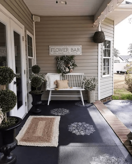 a pretty farmhouse porch with potted trees, a bench with pillows, a wreath with blooms and a basket with greenery