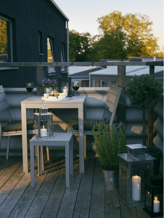 a small and cozy summer balcony with grey wooden furniture, potted greenery and blooms and candle lanterns