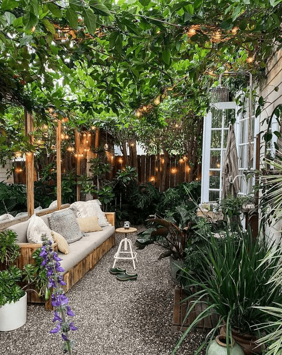 a small and welcoming backyard done with gravel with a sofa, potted plants and blooms, climbing plants over the space and lights