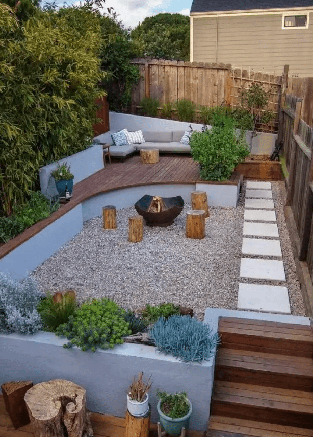 a small and welcoming backyard with a fire pit zone and tree stumps with growing plants and trees, with a raised sitting zone with a sofa