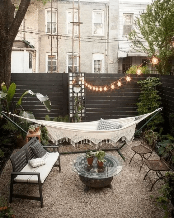 a small backyard with a couple of chairs, a bench and a coffee table, a hammock, some greenery and string lights over the space