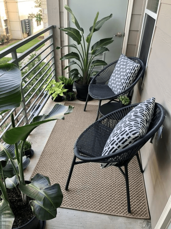 a small modern balcony with a jute rug, black chairs and pillows and some potted tropical plants