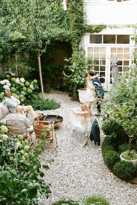 a small yet cozy backyard with gravel on the ground, some rattan furniture and lots of greenery and white blooms around